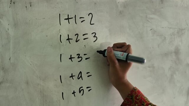 Close up of a hand writing basic math addition equations with a black marker on a whiteboard