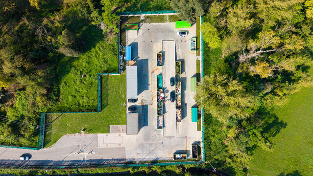 Aerial view of a recyclable waste collection point with various containers and bins arranged in a paved area surrounded by trees in Bettola-Zeloforamagno, Lombardia, Italy.