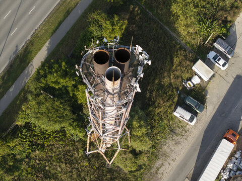 Aerial view of a metal telecommunications tower with three large cylindrical vents at the top, surrounded by green trees, a road, and parked vehicles in Krakow, Lesser Poland Voivode, Poland.