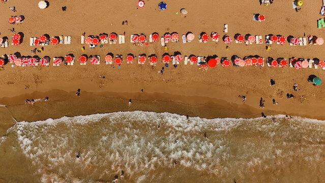 Aerial view of Alanya Incekum beach with rows of red sun umbrellas, sun loungers, and people relaxing on the sand by the turquoise sea in Alanya, Antalya, Turkiye.