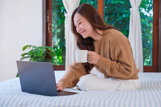 Portrait image of a woman working on laptop computer and drinking coffee while sitting on a bed at home
