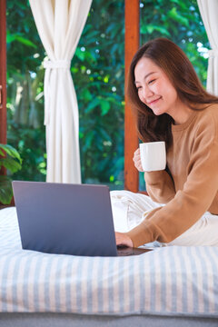 Vertical portrait image of a woman working on laptop computer and drinking coffee while sitting on a bed at home