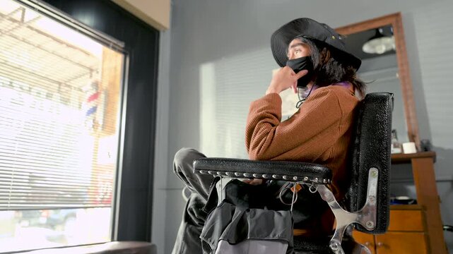 A barber handles a black salon chair beside a seated customer. The barber works near the chair&rsquo;s padded upholstery and metal armrest as soft daylight reaches the barbershop interior.