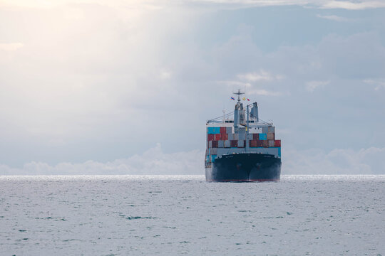 Distant container ship sailing on a calm ocean under a bright, cloudy sky, seen from astern, symbolizing global shipping, freight logistics, and international maritime trade.