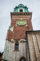 Wawel Cathedral's imposing Sigismund Clock Tower, featuring a detailed green and gold clock face, rich brickwork, and carved stone elements under an overcast sky in Krakow, Poland. © Danica Chang