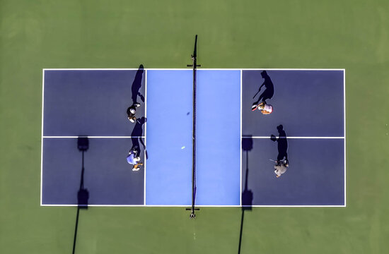 Aerial view of a pickleball game with four players on a blue and dark blue court with long shadows in Valencia, California, United States.