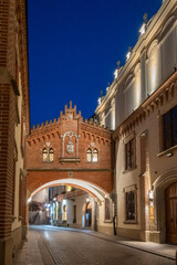 Alley of Princes Czartoryski, a picturesque brick archway connects historic buildings above a narrow cobblestone street, softly illuminated at night in Krakow, Poland. © Danica Chang