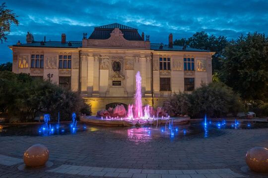 Vibrant pink and blue water jets illuminate a decorative Szczepanski fountain, set against a grand, lit-up Palace of Art. Lush greenery surrounds the nocturnal scene in Krakow, Poland.
