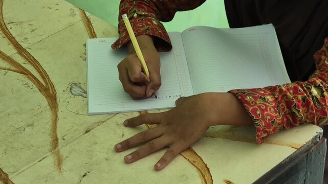 Close up of a person wearing a batik shirt writing in a notebook with a yellow pencil
