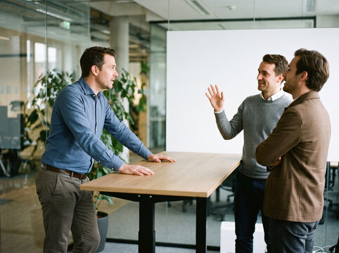 Three male colleagues in candid discussion around standing desk with blank whiteboard