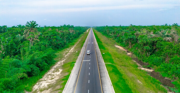 Aerial view of a straight road stretching through the dense tropical forest of Bonny Island with a single vehicle driving along the asphalt surface in Bonny Town, Rivers, Nigeria.