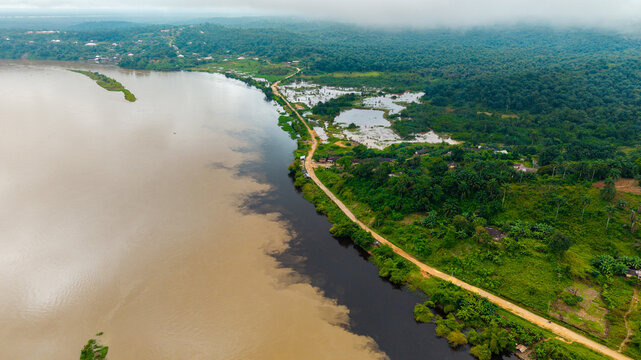 Aerial view of the river confluence near Itu, featuring contrasting water currents, lush tropical vegetation, and a dirt road along the riverbank in Itu, Akwa Ibom, Nigeria.