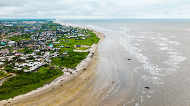 Aerial view of Ibeno Beach featuring a long sandy coastline, small fishing boats in the surf, and the town of Kwa Ibo with green vegetation under a cloudy sky in Kwa Ibo, Akwa Ibom, Nigeria.