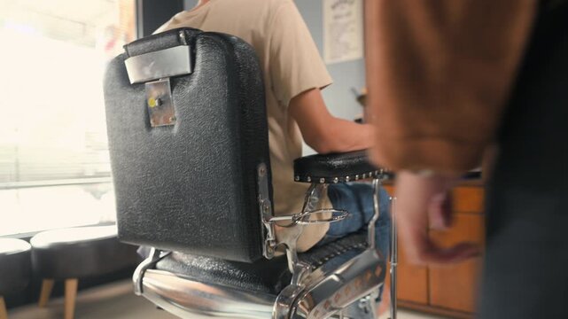 An empty barber chair sits inside a barbershop with black upholstery and a metal armrest. The barbershop interior shows the barber chair near a mirror, cabinet, bright window, and tiled floor.
