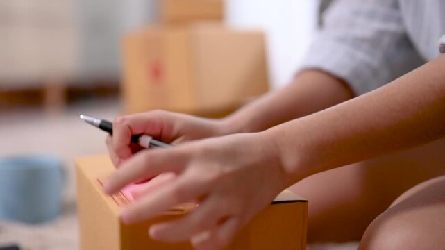 Cardboard boxes of different sizes form a parcel stack inside a home packing area. The cardboard boxes include open and closed brown cartons on the carpet beside a sofa and packing supplies.