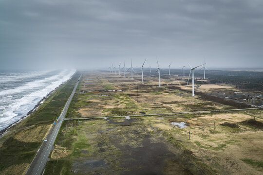 Aerial view of a wind farm with multiple turbines standing along the rugged coastline and a road running parallel to the sea in Wakkanai, Hokkaido, Japan.
