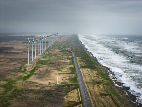 Aerial view of a long row of wind turbines stretching along the coastline, with a road running parallel to the sea in Wakkanai, Hokkaido, Japan.