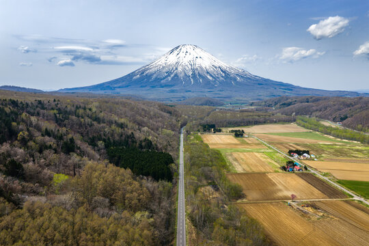 Aerial view of Mount Yotei with its snow-capped peak rising above the surrounding forests, agricultural fields, and a straight road leading towards the horizon in Niseko, Hokkaido, Japan.