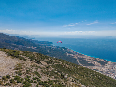 Aerial view of Bregdet.al Llogara Pass with a paraglider soaring above the rugged mountain slopes and the turquoise Ionian Sea coastline in Llogara, Vlore County, Albania.