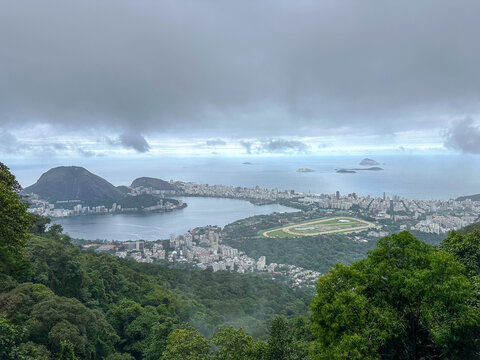 Panoramic scenic view of Rio de Janeiro featuring Rodrigo de Freitas lagoon and Jockey Club surrounded by lush tropical rainforest under dramatic misty overcast sky.