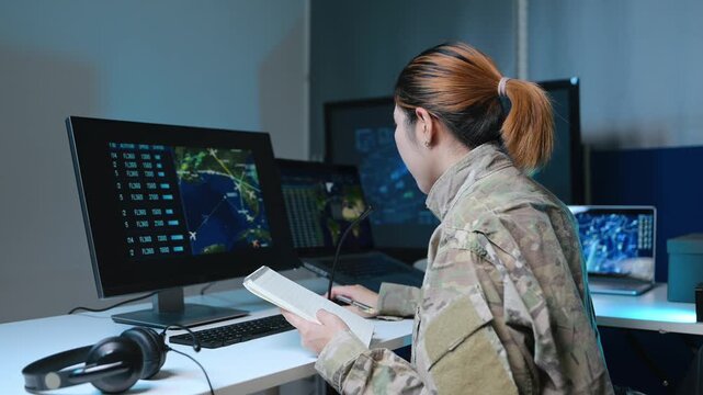 Asian female military officer in camouflage uniform analyzes radar maps on screens and writes notes in a high-tech tactical command center.