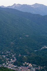 Naklejka premium Houses cluster together at the foot of a lush, wooded mountainside in a deep alpine valley, below rugged terrain. Snowy distant peaks loom above the dense forest canopy beneath a soft hazy evening sky