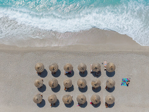 Aerial view of rows of straw umbrellas and sun loungers on a sandy beach with turquoise sea waves washing onto the shore in Albania.
