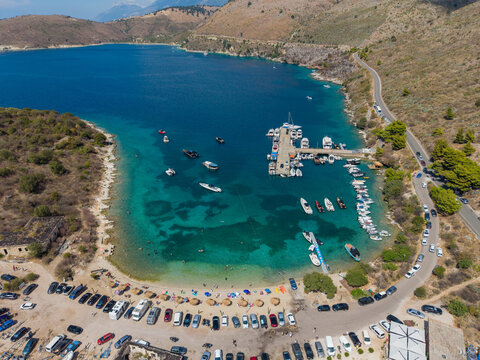 Aerial view of The Castle of Porto Palermo on a peninsula in a turquoise bay with boats at a pier, a sandy beach, and a coastal road in Porto Palermo, Qarku i Vlores, Albania.