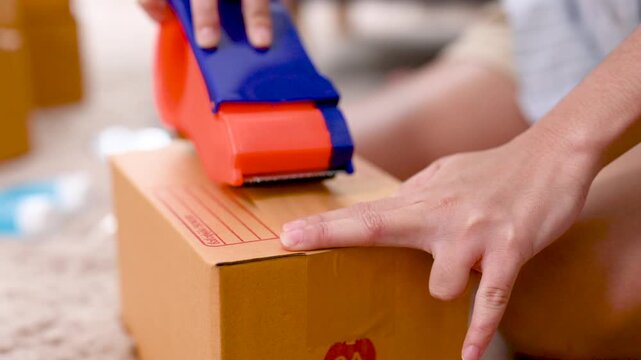 Hands press a tape dispenser against a parcel box to seal the cardboard surface. The hands guide clear packing tape across the box seam as the orange and blue tool moves over the brown carton.