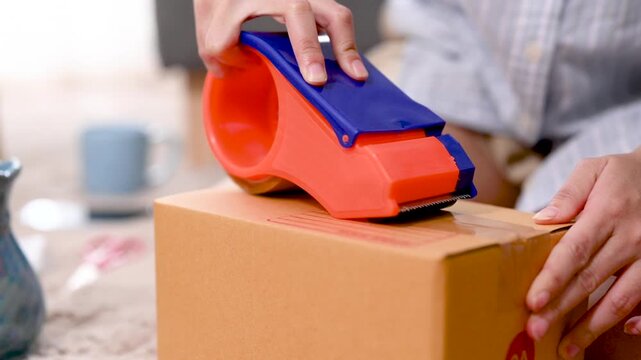 Hands press a tape dispenser against a parcel box to seal the cardboard surface. The hands guide clear packing tape across the box seam as the orange and blue tool moves over the brown carton.