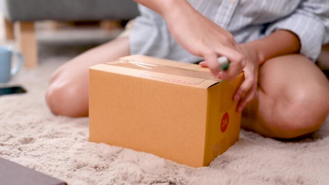 Hands press a tape dispenser against a parcel box to seal the cardboard surface. The hands guide clear packing tape across the box seam as the orange and blue tool moves over the brown carton.