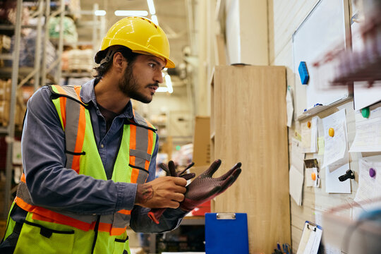 Warehouse worker in hardhat and safety vest reading schedule on notice board while wearing protective gloves in industrial storage facility.