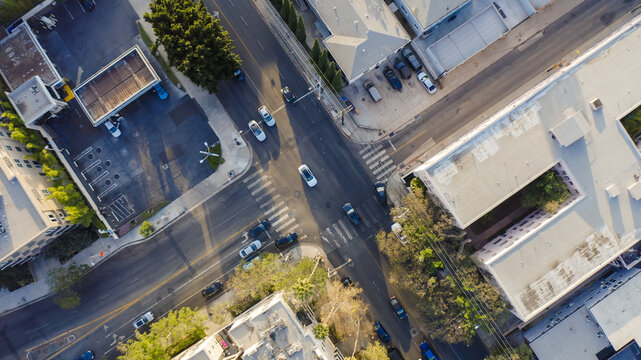 Aerial view of a street intersection with cars driving on asphalt roads, surrounding commercial buildings, parking lots, and green trees in Los Angeles, California, United States.
