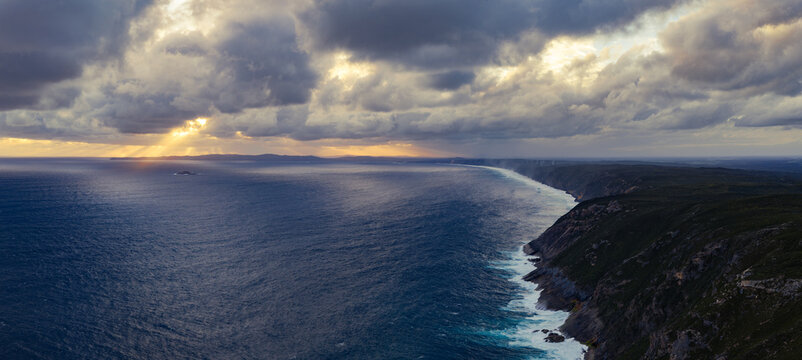 Aerial view of Sharp Point Lookout featuring dramatic cliffs, crashing waves, deep blue ocean, and sun rays breaking through clouds at sunset in Torndirrup, Western Australia, Australia.