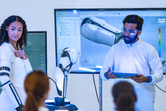 Bearded engineer in goggles gestures toward a robotic arm simulation on screen while curly-haired woman wearing an exoskeleton arm stands beside him, a next generation prosthetics concept