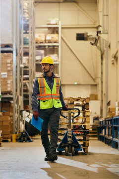 Full length of warehouse worker pulling pallet jack through industrial facility.