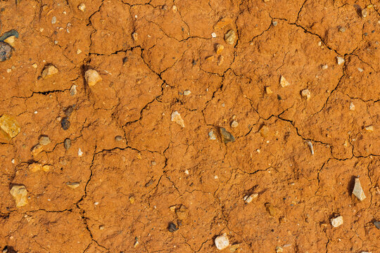 Close-up of parched cracked red earth texture with small scattered stones and gritty soil forming a natural abstract arid desert pattern under warm bright sunlight