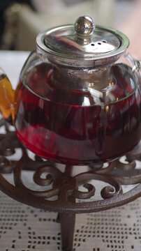 Glass teapot filled with dark red hibiscus tea resting on vintage cast iron trivet over white lace tablecloth