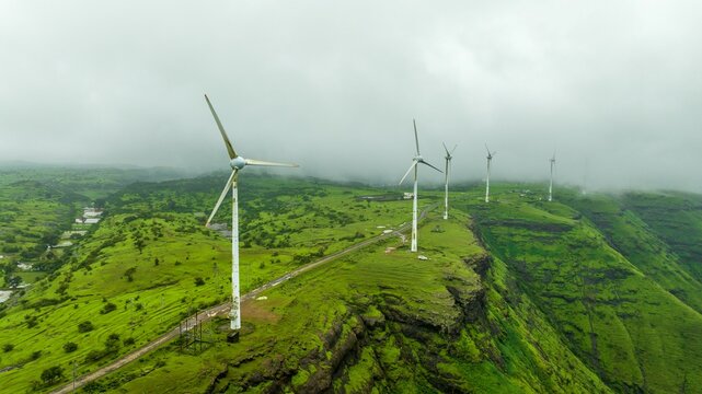 Aerial view of wind turbines standing on a lush green hillside under a misty sky in Igatpuri, Maharashtra, India.