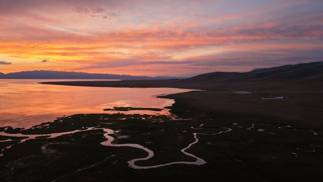 Aerial view of Song-Kul lake at sunset with a winding river inlet, rolling hills, and vibrant orange sky reflecting on the calm water in Naryn Region, Kyrgyzstan.