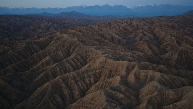Aerial view of the rugged, eroded rock formations of Skazka Canyon with snow-capped mountains in the distance under a twilight sky in Issyk-Kul Region, Kyrgyzstan.