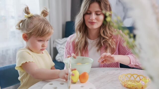 Young girl pours orange liquid into cup, assisted by smiling mother. Mother watches with gentle expression, guiding child's hand. Warm, bright kitchen setting with Easter eggs and basket