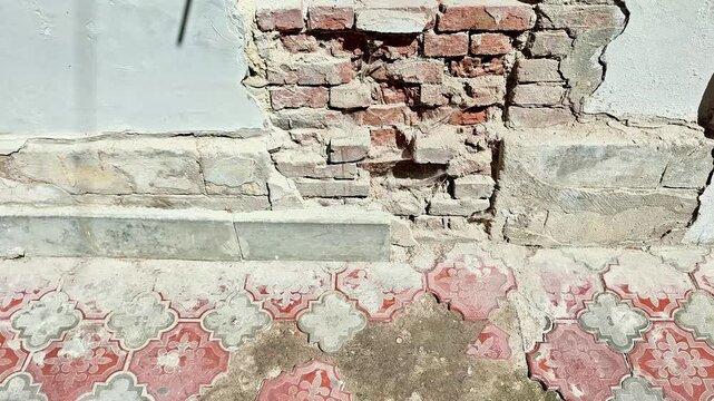 Damaged interior wall with exposed brickwork, cracked plaster, weathered surfaces, and patterned flooring, showing deteriorated building materials and unfinished renovation