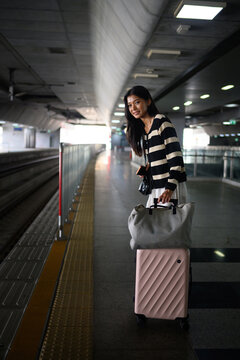 Young Asian Woman Traveler Standing on Train Platform with Pink Suitcase and Tote Bag Smiling
