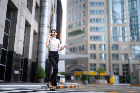 Young Businesswoman Walking in City While Talking on Smartphone and Carrying Laptop