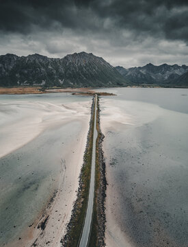 Aerial view of Midnattsolveien road cutting through shallow tidal flats with dramatic mountains in the background under a cloudy sky in Laukvik, Nordland, Norway.