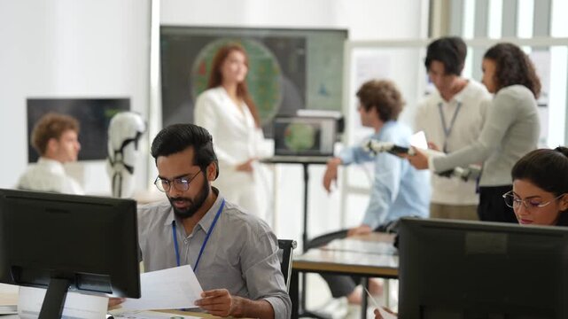 Bearded engineer reviews papers at his desk while teammates test a bionic hand and discuss screens in a busy tech office, during an iterative design check, a tech startup innovation concept