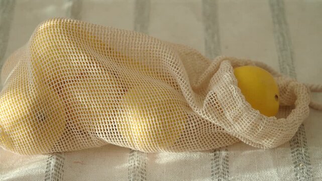 A close up view of several fresh, organic lemons in a reusable mesh bag, lying on a white cloth, captured with smooth camera motion