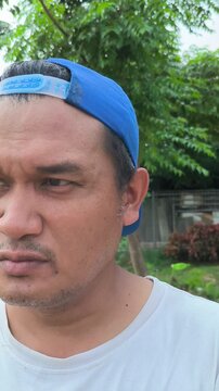 Vertical selfie video of an Asian man wearing a backward blue cap and white t-shirt looking around with a serious expression in an outdoor park setting