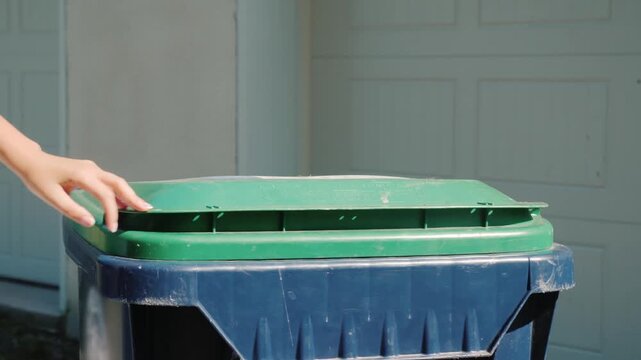 Person disposing black plastic trash bag into blue recycling bin with green lid outside residential garage door during sunny daylight hours showing close up action of waste management daily 